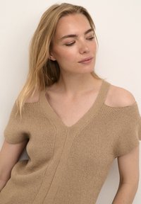 Haut tricoté marron clair avec des manches courtes et un large col en V. Présente des découpes ouvertes aux épaules et des lignes texturées qui courent verticalement.
