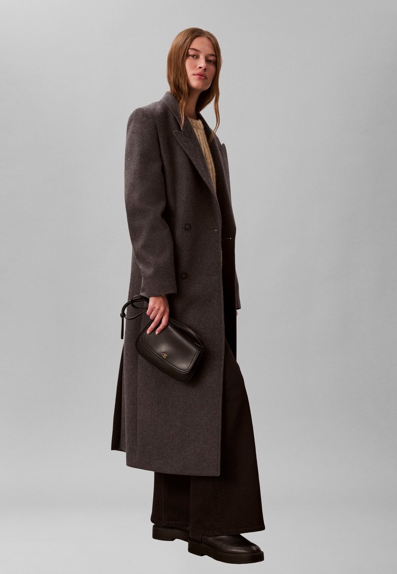 Femeie stă în picioare purtând o haină lungă gri cu nasturi dubli, pulover bej, pantaloni largi negri, cizme negre, ținând o geantă mică neagră pe o parte.