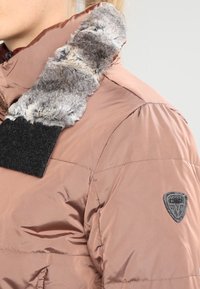 Chaqueta acolchada de material beige suave con un cuello de piel sintética gris, parche de velcro negro y una placa con el logo en gris en la manga.