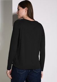 Musta pitkähihaine paita, jossa on pyöreä pääntie ja teksturoidut olkatyynyt, yhdistetty tummansinisihin farkkuihin. Materiaali näyttää pehmeältä ja sileältä.