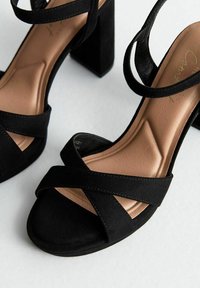 Sandalias de tacón alto de ante negro con correas cruzadas, tacón cuadrado y plantilla beige lisa con un diseño moldeado.