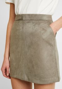 Suede mini sukně v tlumené olivové barvě, s vysokým pasem a postranními kapsami. Měkká textura a rovný střih s minimálními švy.