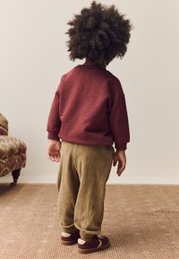 Enfant portant un sweat-shirt bordeaux, un pantalon en velours côtelé vert olive et des baskets marron avec des accents beige. Vue de dos, montrant des cheveux bouclés.