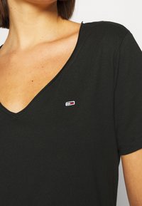 Žena v černém tričku s výstřihem do V, na prsou má malé obdélníkové logo v červené, bílé a modré barvě, tričko končí pod rameny.