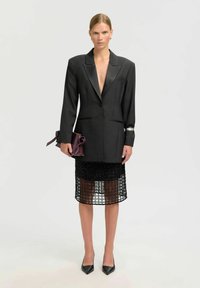 Ženska v prevelikem črnem blazerju, črni mrežasti krilu, črnih petah, drži vijolično torbico in stoji pred enostavno svetlo ozadjem.