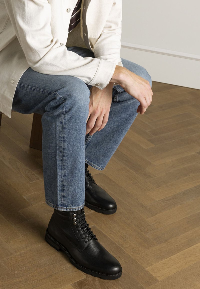 Bottes en cuir noir avec un design à lacets et un bout rond, portées avec un jean en denim bleu clair et une chemise en coton crème, assis sur un sol en bois.