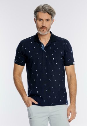 Mężczyzna z siwymi włosami i brodą, ubrany w granatową koszulkę polo w drobne białe wzory i jasne spodnie, stoi z jedną ręką w kieszeni.