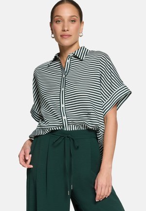 Femme portant une chemise à manches courtes à rayures horizontales noires et blanches, boutonnée et rentrée dans un pantalon vert foncé à cordon, se tenant devant un fond uni.