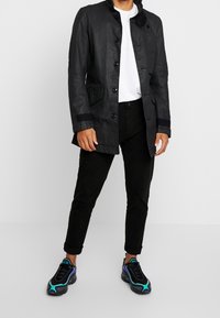 Cappotto lungo in pelle nera con frontale con bottoni, tasche laterali e colletto alto. Abbinato a pantaloni neri e sneakers multicolori distintive.