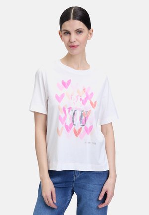 T-shirt blanc à manches courtes avec un imprimé de cœur coloré dans des teintes de rose et de pêche, portant le texte "SOIS TOI" en argent, coupe décontractée.