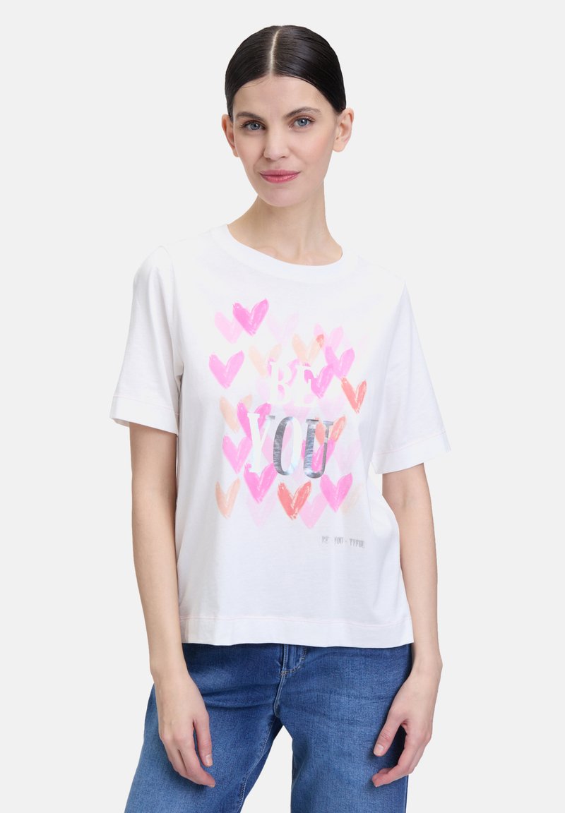 T-shirt blanc à manches courtes avec un imprimé de cœur coloré dans des teintes de rose et de pêche, portant le texte "SOIS TOI" en argent, coupe décontractée.