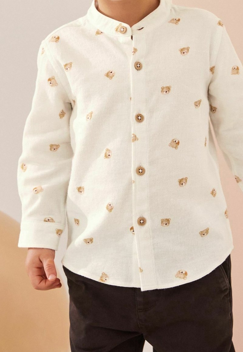 Enfant portant une chemise blanche à boutons avec un motif de visages d’ours en peluche et un pantalon foncé, debout devant un fond neutre.