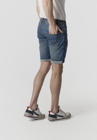 Egy férfi, aki felhajtott farmer rövidnadrágot és fehér tornacipőt visel, egyik kezét a hátsó zsebében tartva áll egy egyszerű háttér előtt.