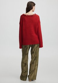 Roter, übergroßer Strickpullover mit strukturiertem Stoff und einer Kettenakzentuierung am hinteren Halsauschnitt, kombiniert mit olivgrünen, gemusterten, weit geschnittenen Hosen.