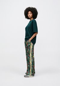 Grøn t-shirt i bomuld kombineret med bukser med blomsterprint og gule striber. Modellen har grå sko på og krøllet hår.