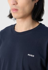 Tamsiai mėlynas medvilninis marškinėliai su apvaliu kaklu, ant kairės krūtinės yra mažas baltas "HUGO" logotipas. Minkšta tekstūra, minimalus dizainas.