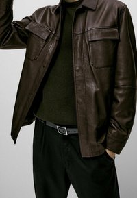 Ruskea nahkainen kauluspaita, jossa kaksi rintataskua, päällä tummanvihreä neulepusero. Mustat housut ja vyö täydentävät asun.