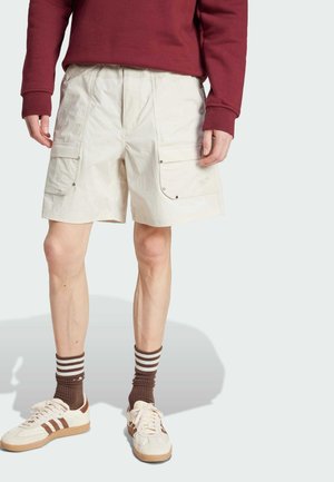 Beige cargo shorts with side pockets, adjustable waistband, and slight texture. Paired with a maroon long-sleeve shirt and beige sneakers.