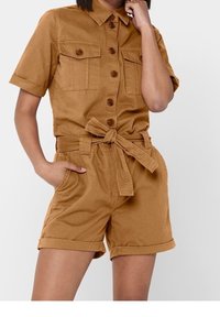 Combinaison courte en coton marron à manches courtes avec fermeture boutonnée sur le devant, deux poches poitrine, ceinture à la taille et shorts retroussés. Texture lisse.