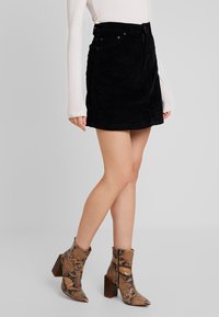 Zwarte corduroy mini-jurk met voorzakken, gecombineerd met tan slangenprint enkellaarzen met een blokhak. Eenvoudig wit shirt met lange mouwen.