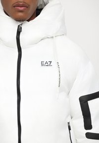 Biela nafukovacia bunda s čiernym zipsom a veľkým grafickým logom na rukáve. Obsahuje vysoký golier a kapucňu so sťahovacou šnúrkou.