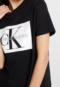 Schwarzes Baumwoll-T-Shirt mit einem weißen rechteckigen Grafik, auf der "CK" in fetten schwarzen Buchstaben und "CALVIN KLEIN JEANS" in Grau steht, mit kurzen Ärmeln.