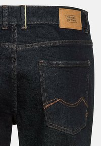 Ciemnoniebieskie dżinsy z denimu z skórzaną naszywką Camel Active na pasku, kontrastowymi pomarańczowymi szwami oraz dwiema tylnymi kieszeniami z zakrzywionymi szwami.