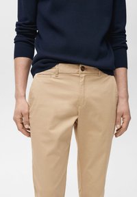 Bež chinos hlače z gladko teksturo, ki imajo gumb za zapiranje in stranske žepe, v kombinaciji z mornarsko modrim pletenim puloverjem.