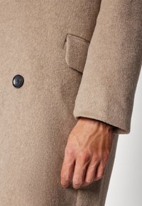Beiger Wollmantel mit strukturiertem Finish, einem schwarzen Knopf und einer Seitentasche. Der Ärmel hat ein umgeschlagenes Detail. Hand liegt in der Nähe.