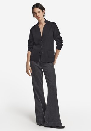 Schwarze Strickjacke mit Stehkragen, Knopfverschluss, gerippten Bündchen und zwei vorderen Taschen, kombiniert mit dunkelblauen weiten Jeans.