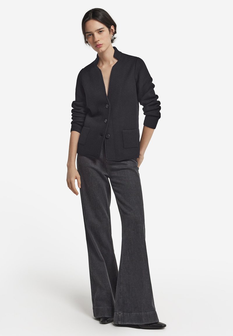 Svart stickad cardigan med ståkrage, knäppning, ribbade muddar och två framfickor, tillsammans med mörka vida jeans.