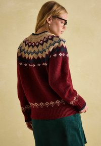 Roter gestrickter Pullover mit gemustertem Einsatz in Creme und Marineblau; gerippte Bündchen und Saum; weiche Textur; über einem grünen Cordrock getragen.