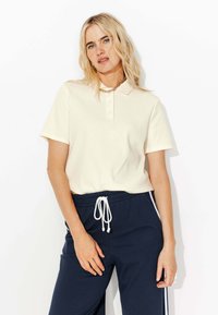 Polo beige avec un col et des manches courtes, associé à un pantalon de jogging bleu marine avec un cordon de serrage blanc et des bandes latérales.