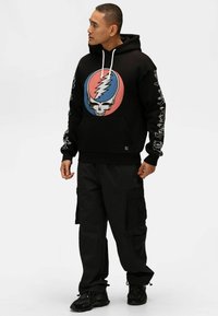 Felpa nera con cappuccio presenta una grande grafica di teschio colorato sul davanti, lacci bianchi e disegni intricati sulle maniche. Indossata con pantaloni cargo.