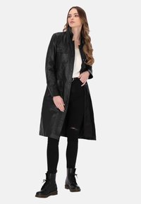 Mustahi leather coat, jossa on napitettava etuosa, pitkät hihat ja kaksi rintataskua. Päällä beigellä paidalla ja mustilla farkuilla, yhdistetty mustiin saappaisiin.