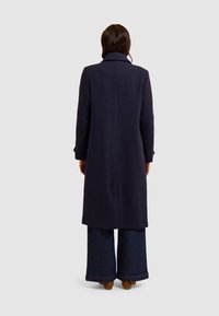 Cappotto di lana blu navy con silhouette tailored, colletto alto e maniche lunghe. Sul retro presenta una cucitura centrale e una texture liscia, abbinato a jeans larghi.