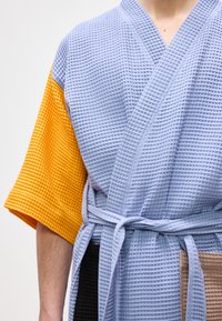 Άτομο που φοράει ρόμπα με υφή πλεκτού βαφλού με ανοιχτό μπλε σώμα, κίτρινο αριστερό μανίκι, μαύρα και μπεζ πάνελ, δεμένη στη μέση.