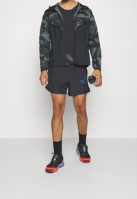 Zwarte sportjack met patroonmouwen, zwart shirt, zwarte shorts met een kleurnuance, en opvallende sneakers met een waterfles.