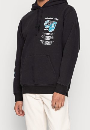 Sweat à capuche noir avec poche avant, texte graphique blanc et bleu « Re-Imagined Living » et design abstrait sur la poitrine, porté avec un pantalon clair.