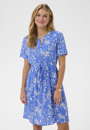 Žena s dugom plavom kosom, odjevena u plavu haljinu kratkih rukava s bijelim cvjetnim uzorcima, stoji pred jednostavnom svijetlom pozadinom.