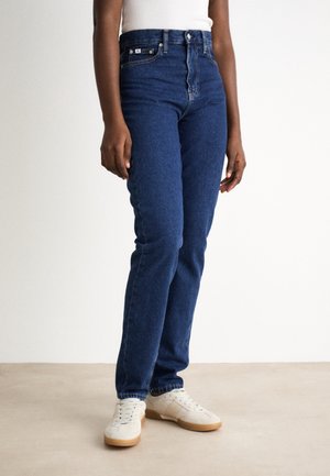 Osoba stojící, oblečená v tmavě modrých džínách s vysokým pasem, bílých teniskách a bílém tílku na jednoduchém pozadí.