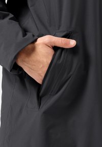 Giacca nera impermeabile con una mano nella tasca laterale, che mostra una texture del tessuto liscia e un design elegante con cuciture minime.