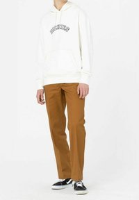 Vit huvtröja med "DICKIES"-logotyp i marinblå, kombinerad med bruna byxor och svarta sneakers med vita sulor.