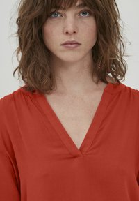 Blusa rossa, morbida e ampia, con scollatura a V e maniche corte. Texture liscia, tessuto leggero e design minimalista.