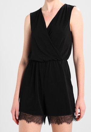 Combinaison noire sans manches avec décolleté en V croisé et bordure en dentelle à l'ourlet, portée par une personne debout devant un fond uni.