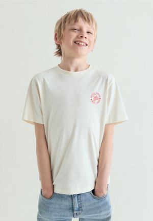 Chico rubio sonriente con una camiseta blanca con logo rojo y vaqueros azules, con las manos en los bolsillos, de pie frente a un fondo claro y liso.