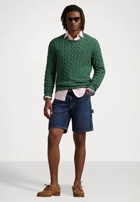 Zaļš džemperis ar rakstu virs gaiši rozā krekla ar apkakli, valkāts kopā ar džinsa šortiem un brūnām zamšādas kurpēm. Aksesuāri: tumši saulesbrilles.