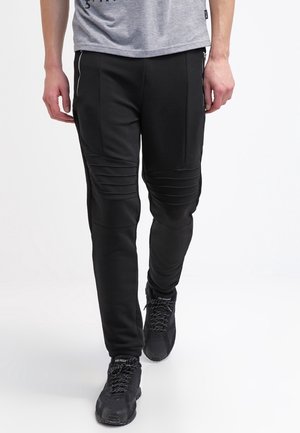 Mann trägt schwarze Bikerhose mit gerippten Kniedetails und schwarzen Sneakers und geht vor weißem Hintergrund.