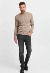 Beige kudumisega sviiter ümmarguse kaelusega ja väikese triibulise logoga rinnal; koos saledate tumesiniste teksade ja mustade kingadega.