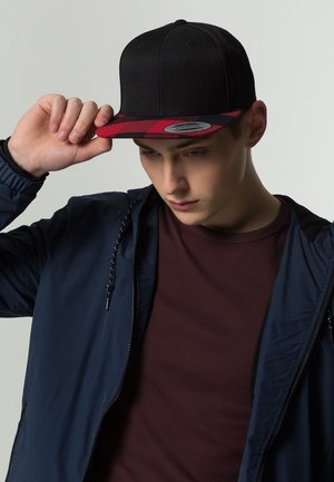 Black snapback cap featuring a red plaid visor. The cap has a textured fabric and a flat brim, with a logo sticker on the front.